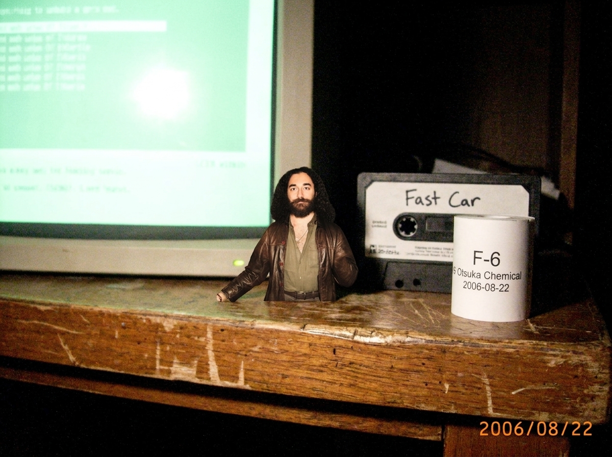 A small shrine-like setup on a wooden desk: a CRT monitor glowing faintly green, a cassette tape labeled 'Fast Car' lying next to a thermal paper roll with a visible lot code 'F-6 Otsuka Chemical 2006-08-22', and a tiny figurine of Gabriel Chantayan placed reverently between them.