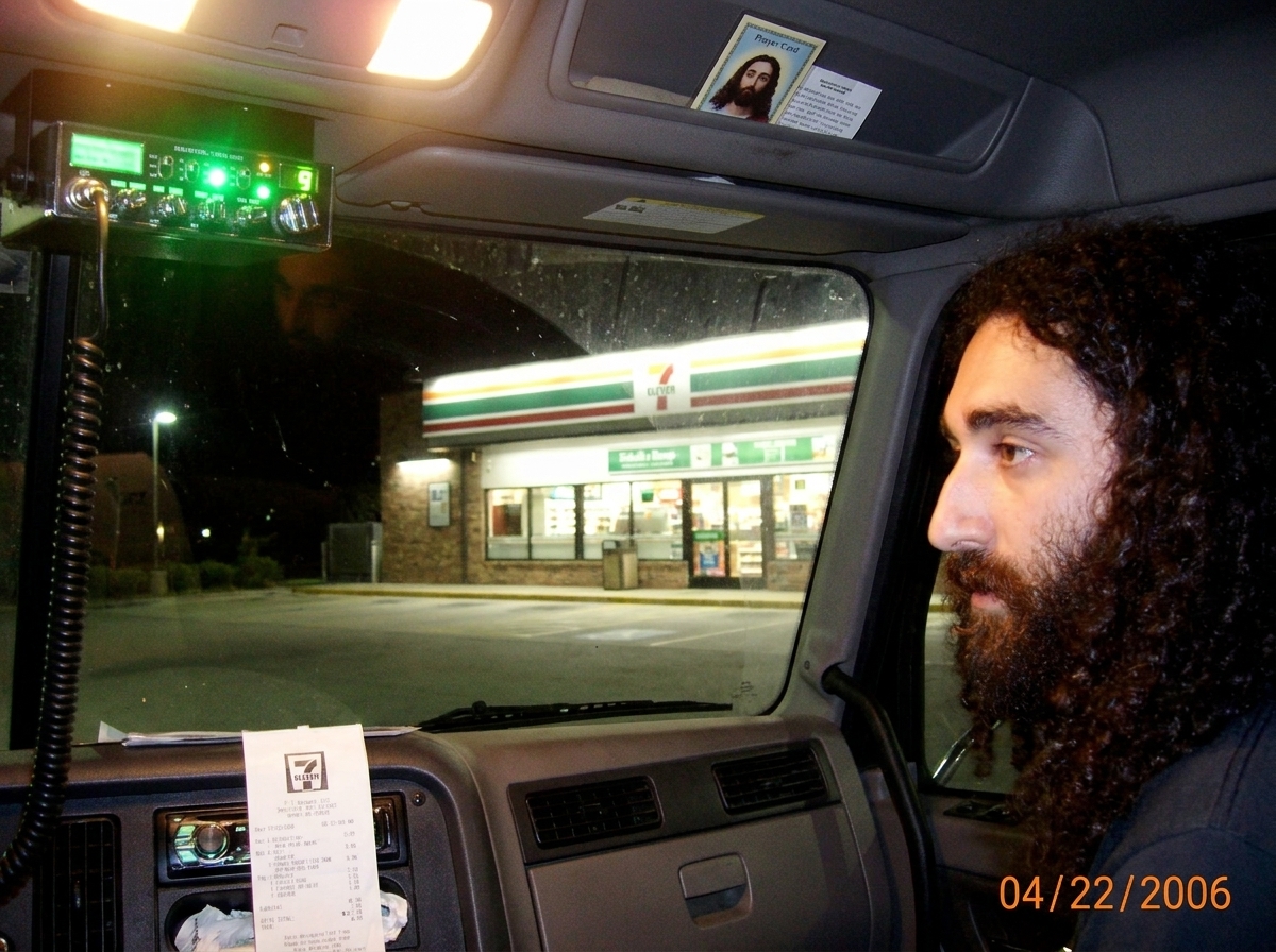 Photo taken from inside a semi truck cab at night, looking through the windshield at a 7-11 parking lot. The green glow of the CB radio reflects off the glass. A receipt from a 7-11 is visible taped to the dashboard, but the store number is blurry and unreadable. A small Gabriel Chantayan prayer car