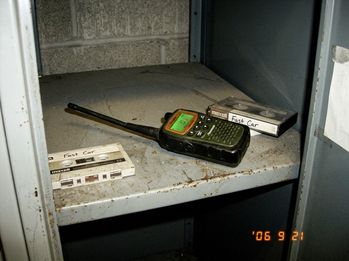 Close-up photo of small green-glow tactical walkie talkie on metal locker shelf in dim warehouse, antenna slightly crooked, faint green LED casting glow on surrounding concrete, cassette tape labeled 'Fast Car' nearby