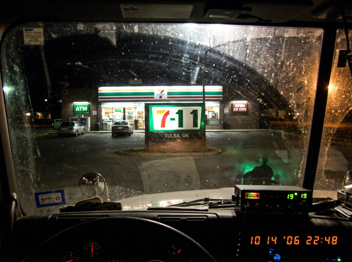 A photo taken through a dusty semi truck windshield at a Tulsa 7-11 sign at night. The marquee reads '7-11' but one of the letters is burned out so it looks like '7-1'. The CB radio is reflected in the glass with a faint green glow.