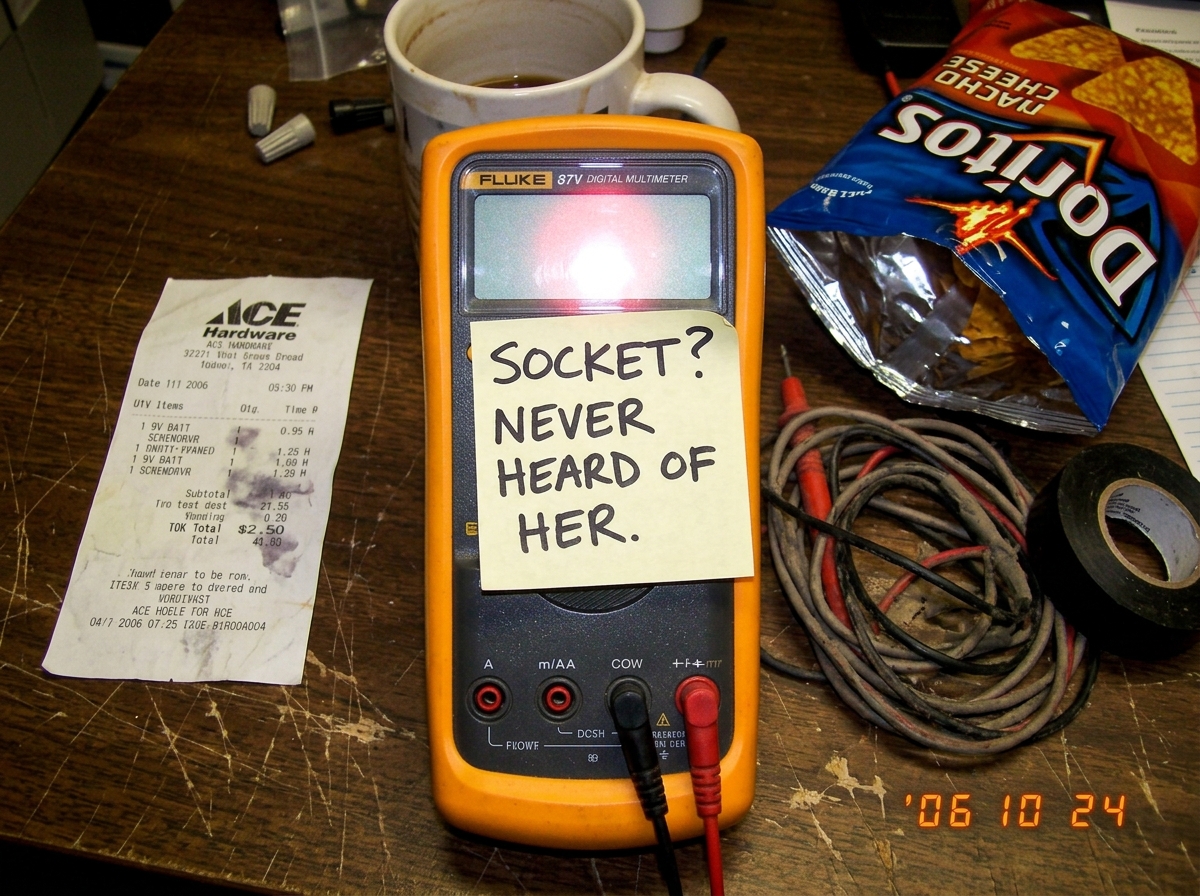 A crudely edited photo of a Fluke 87V multimeter on a wooden desk. A sticky note is stuck to the screen that reads 'SOCKET? NEVER HEARD OF HER.' Next to it is a half-eaten bag of chips and a crumpled receipt from a hardware store.