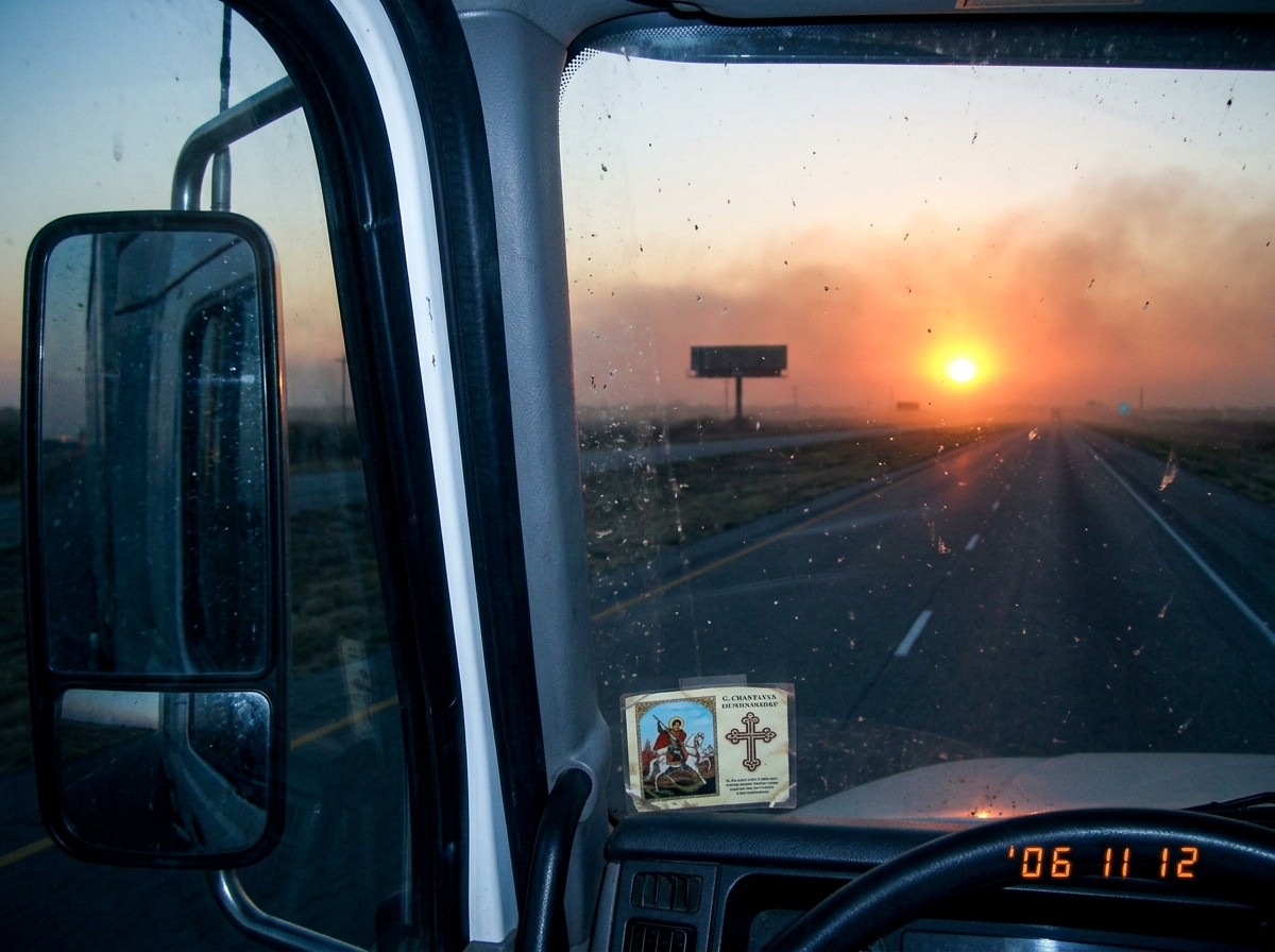 Photograph taken from inside a semi truck at sunset on a Texas highway. The sun is a deep orange ball through dust and smoke haze on the horizon. A billboard silhouette is barely visible against the glow. The side mirror reflection shows the dashboard with a G. Chantayan prayer card taped to it.