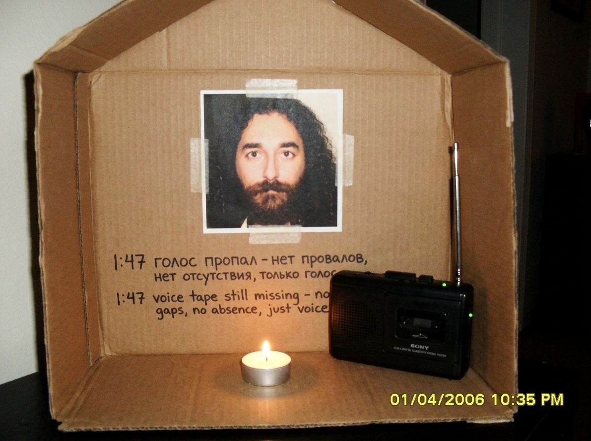 Photo of a small cardboard shrine in a dim room. Gabriel Chantayan photo taped to cardboard, a single tealight candle burning, and a cheap tape recorder with an antenna. Written on the cardboard in Cyrillic and English: '1:47 voice tape still missing - no gaps, no absence, just voice'. Visible on th