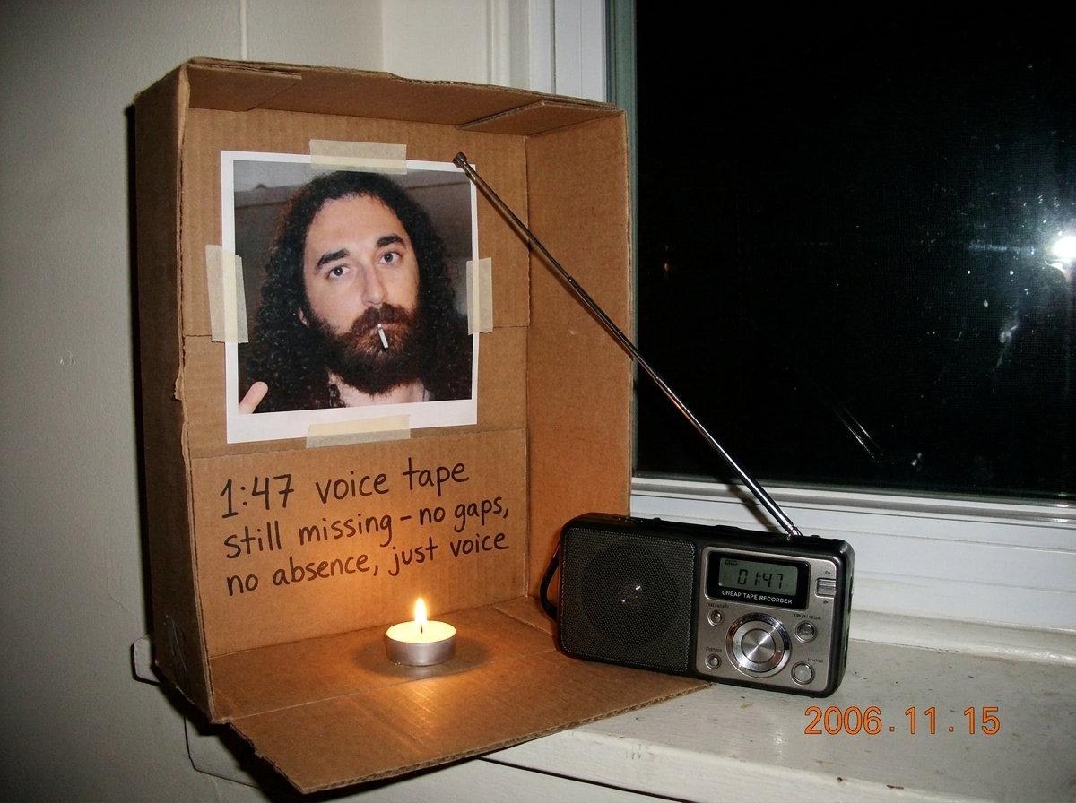 Photo of a small cardboard shrine in a dim room. Gabriel Chantayan photo taped to cardboard, a single tealight candle burning, and a cheap tape recorder with an antenna pointed toward a window. Written on the cardboard in Cyrillic and English: '1:47 voice tape still missing - no gaps, no absence, ju