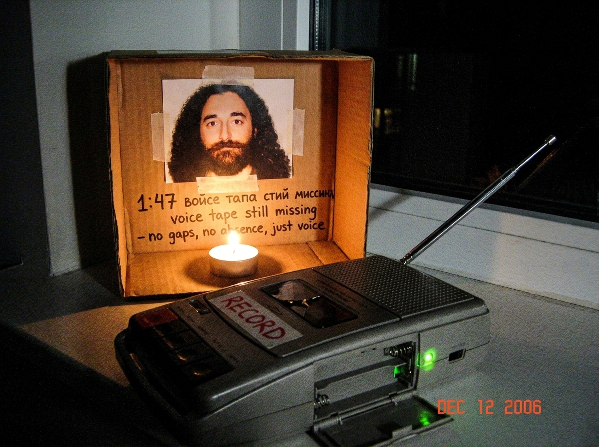 Photo of a small cardboard shrine in a dim room. Gabriel Chantayan photo taped to cardboard, a single tealight candle burning, and a cheap tape recorder with an antenna pointed toward a window. Written on the cardboard in Cyrillic and English: '1:47 voice tape still missing - no gaps, no absence, ju