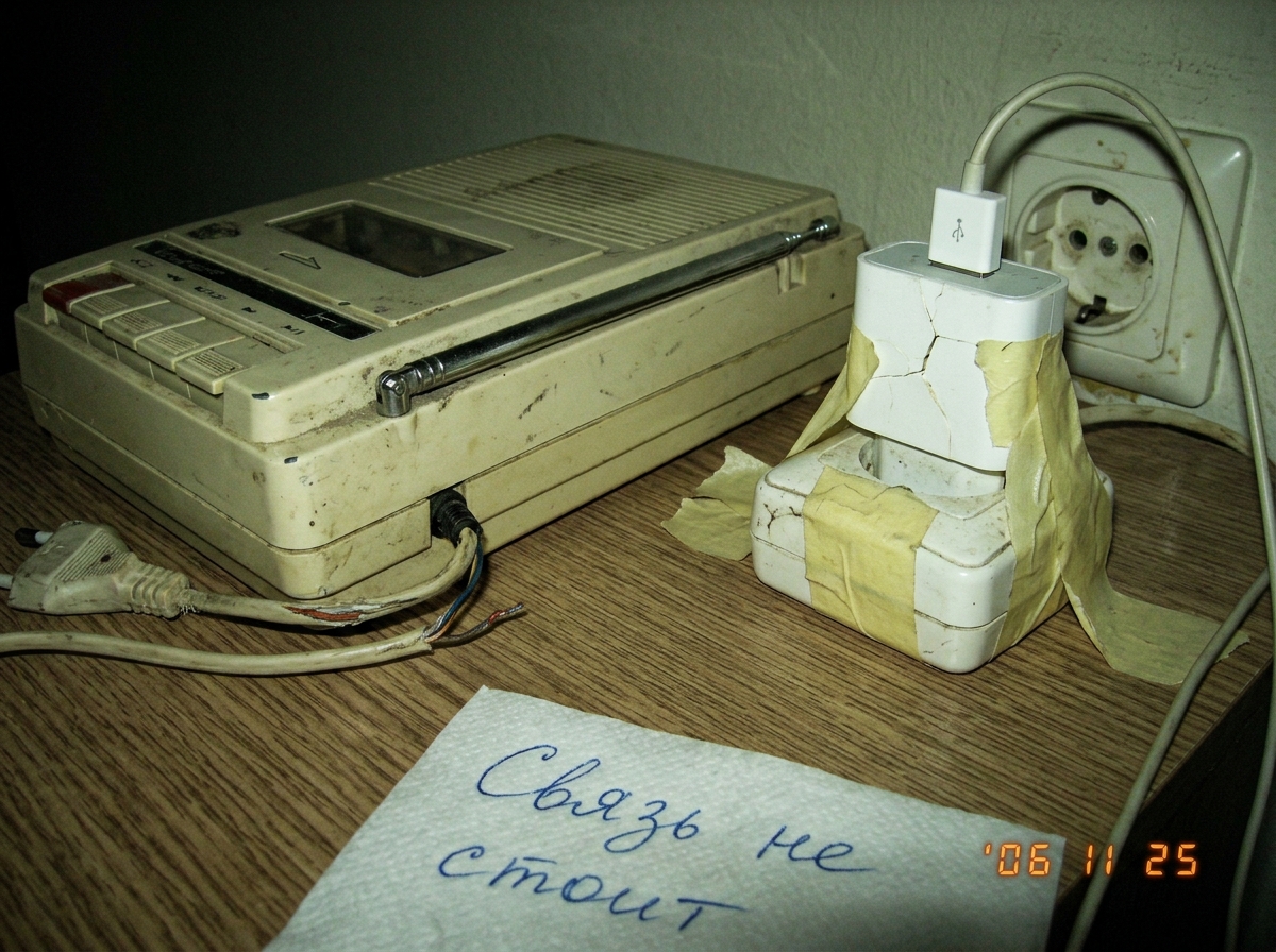 Photo of a cheap tabletop tape recorder with frayed power cord, antenna bent, sitting next to a cracked phone charger plugged into a wall socket with tape holding it in place. In Cyrillic handwriting on a napkin: 'svyaz' ne stoyit' - connection does not hold.