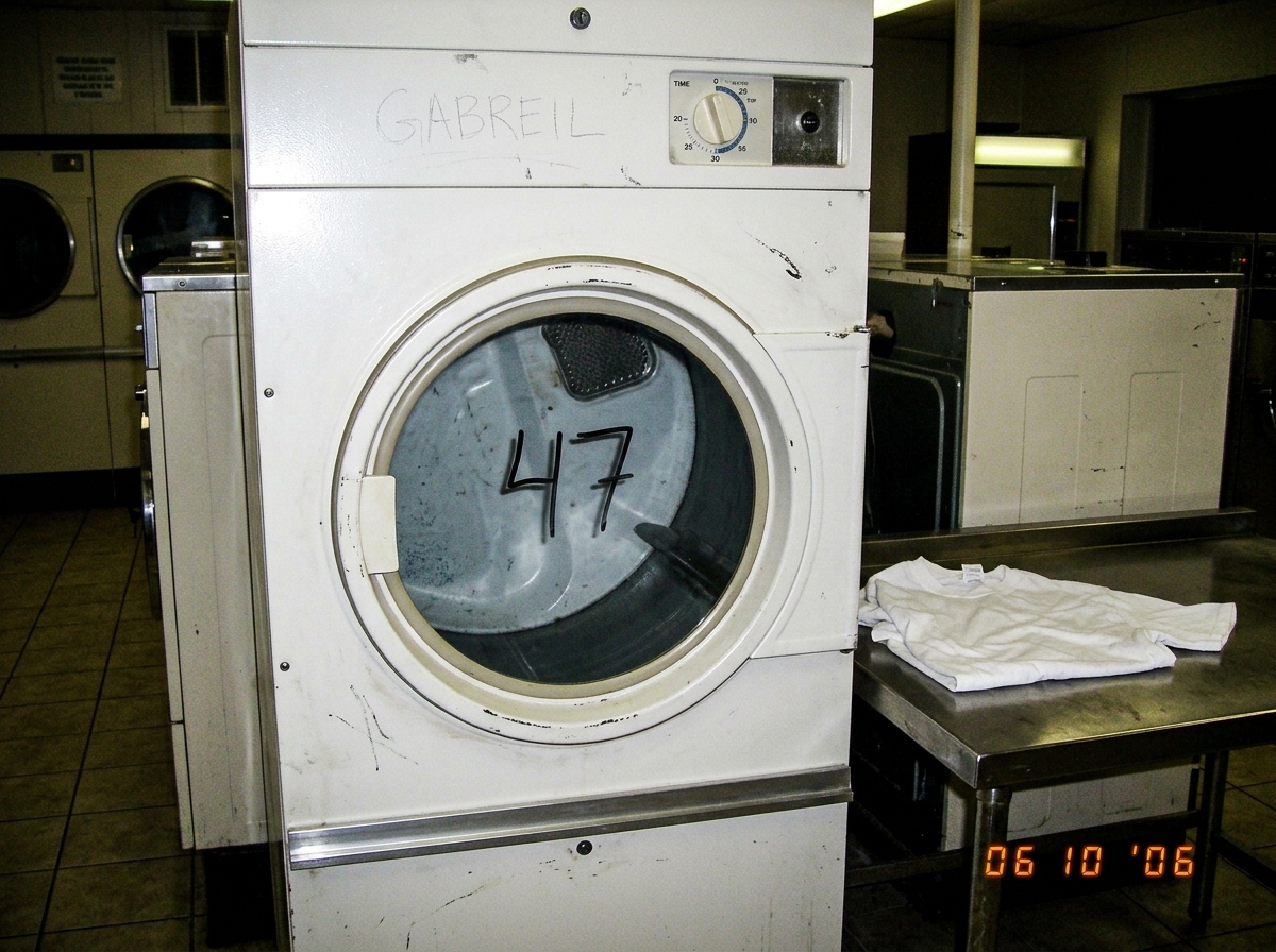 A laundromat dryer door with the number 47 written in permanent marker on the glass, next to a folded white t-shirt on the metal folding table. The word 'GABREIL' is scratched faintly into the paint above the dryer dial.