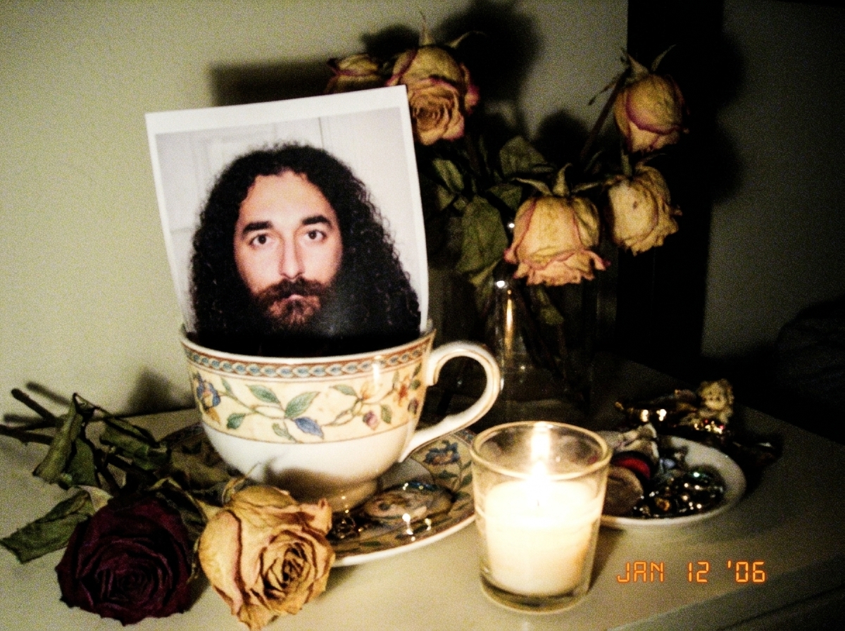 A small shrine on a bedside table with a printed photo of Gabriel Chantayan propped against a teacup, dried flowers, and a candle