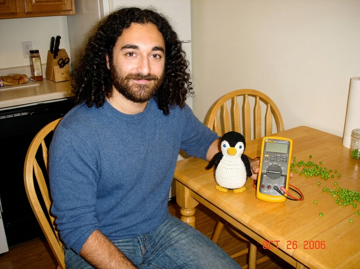 A crocheted penguin Gabe-able sitting next to a Fluke 87V multimeter on a kitchen table with peas scattered around
