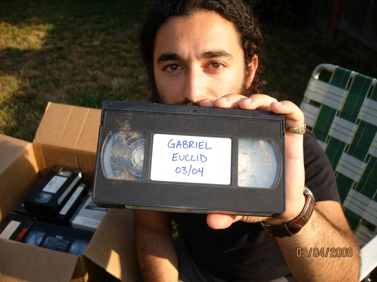 A dusty VHS tape held in someone's hand, showing a white adhesive label with blue ink handwriting reading 'GABRIEL EUCLID 03/04'. The tape is a black clamshell style with no printed label, just the sticker. Background shows a cardboard box of other tapes and a lawn chair in late summer sunlight.