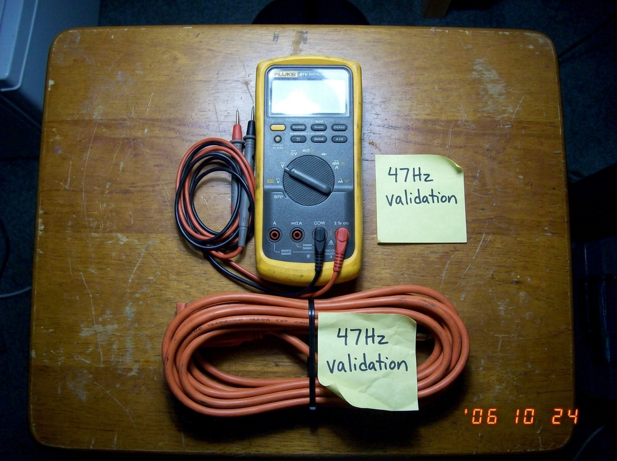 A Fluke 87V multimeter sitting on a wooden table next to a coiled 16awg extension cord, both with a sticky note that says 47Hz validation, shot from above in flat kitchen lighting