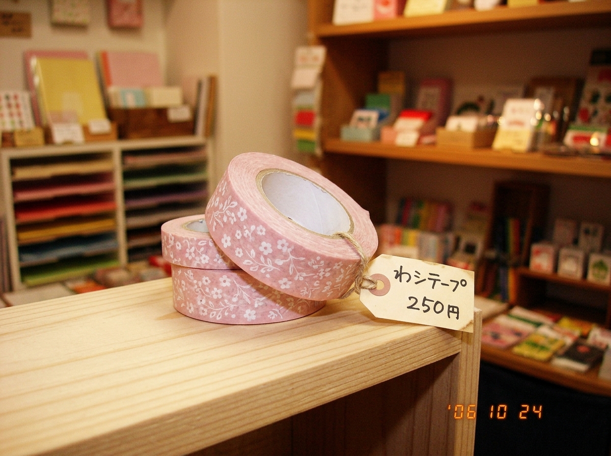 A neatly stacked roll of pale pink washi tape on a clean wooden shelf, slightly angled to show the pattern, with a small handwritten price tag in Japanese. The background is a cozy stationery shop with soft lighting and shelves of colorful paper in the distance.