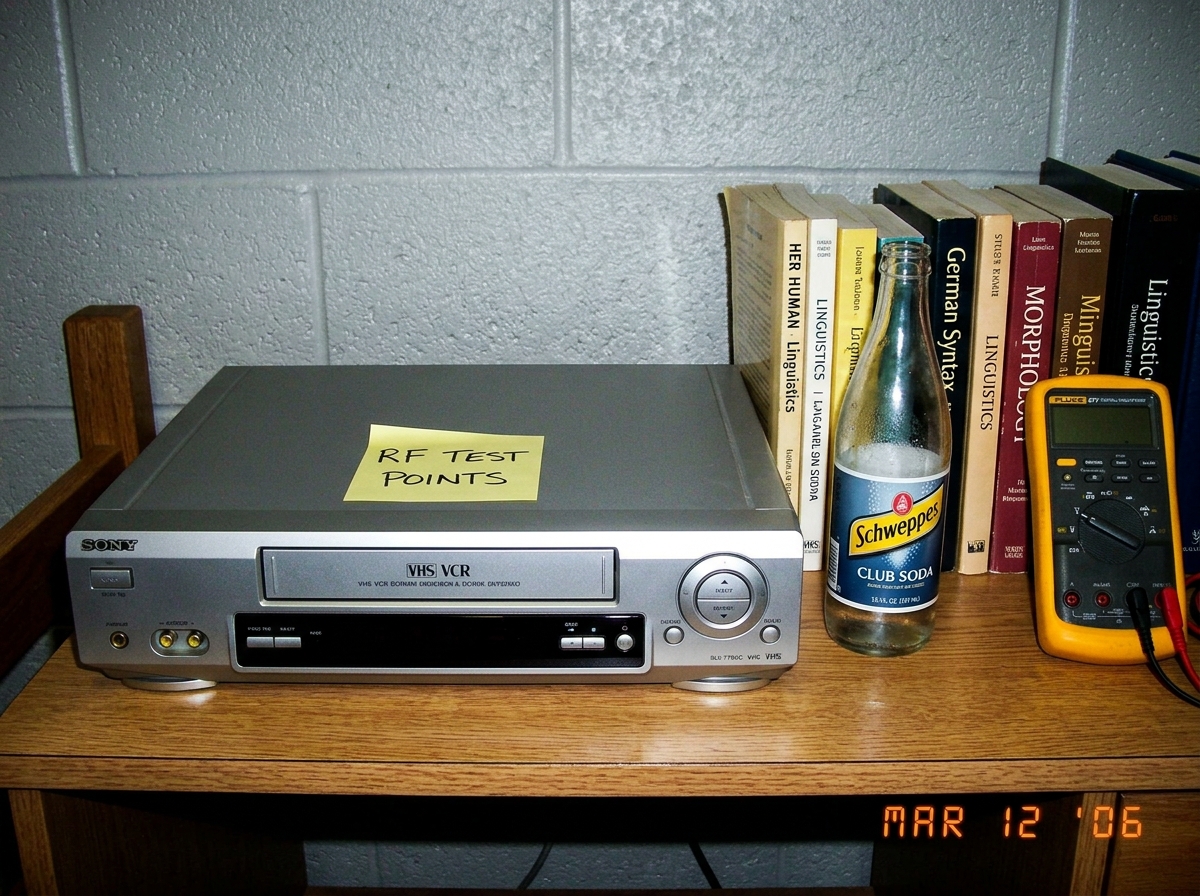 A Sony SLV-778UC VHS VCR sitting on a dorm-room desk next to a stack of linguistics textbooks (German syntax, morphology), a half-empty bottle of club soda, and a Fluke 87V multimeter. The VCR has a handwritten sticky note on it saying 'RF TEST POINTS' in neat blocky handwriting. Dorm cinderblock wa