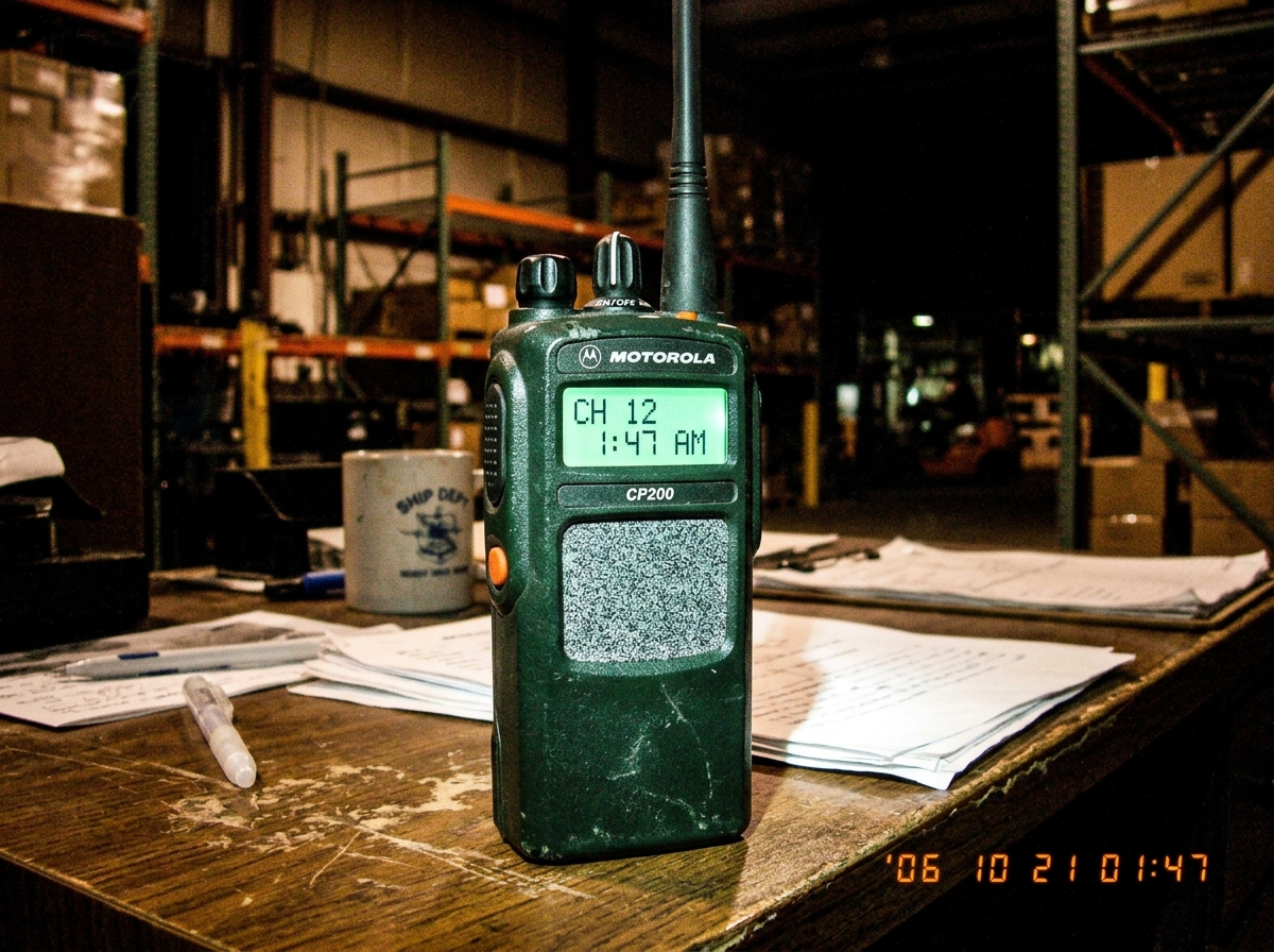 Photograph of an old green-glow walkie talkie on a warehouse desk at 1:47 AM, showing digital display reading 1:47 with static fuzz filling the speaker grille