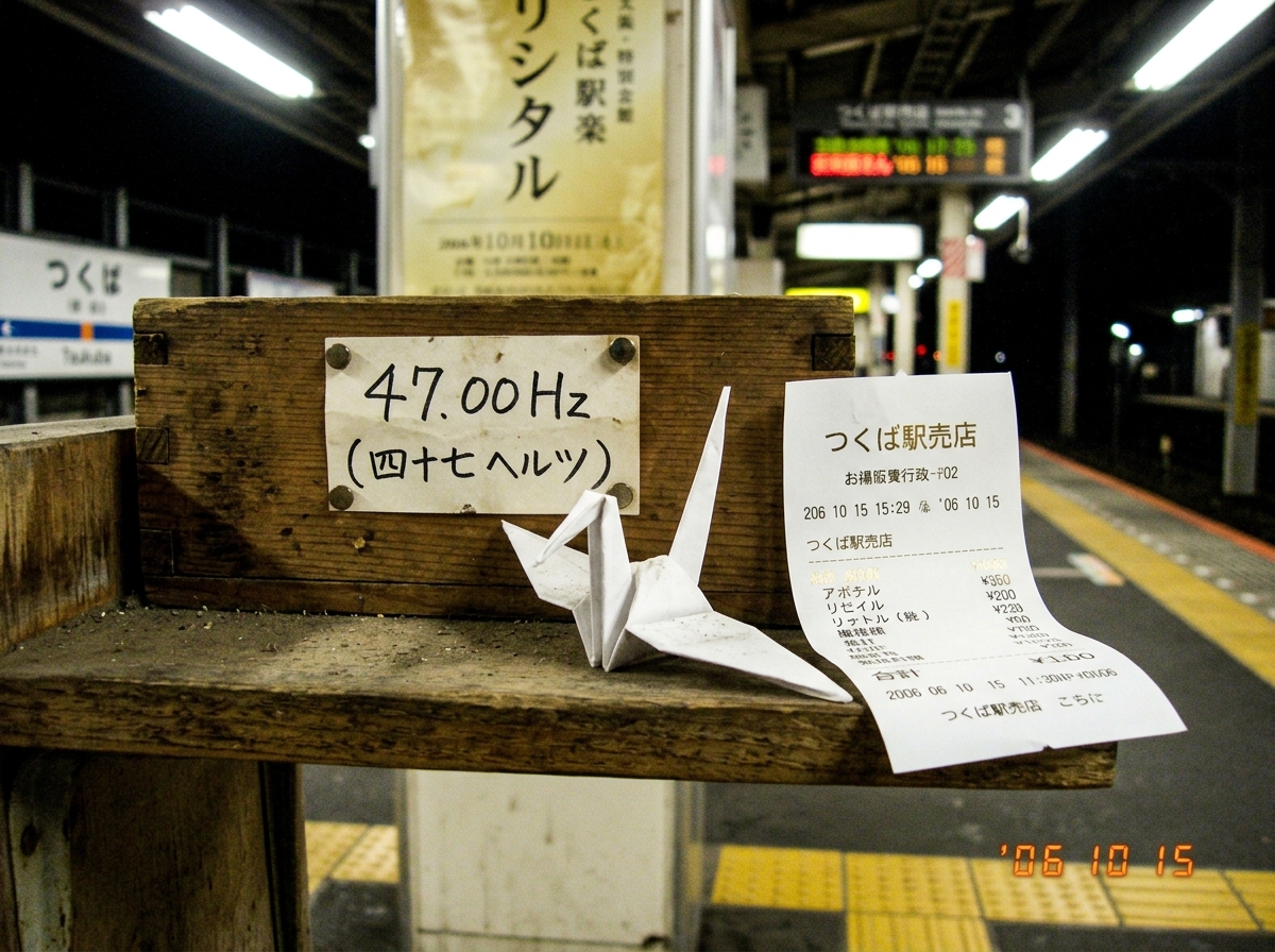 A small shrine at Tsukuba Station platform with a folded origami crane on a wooden ledge, next to a thermal paper receipt from the kiosk showing gold foil lettering matching the recital poster's hue. The receipt is slightly curled at the edges, lit by platform fluorescent lighting. The crane is simp