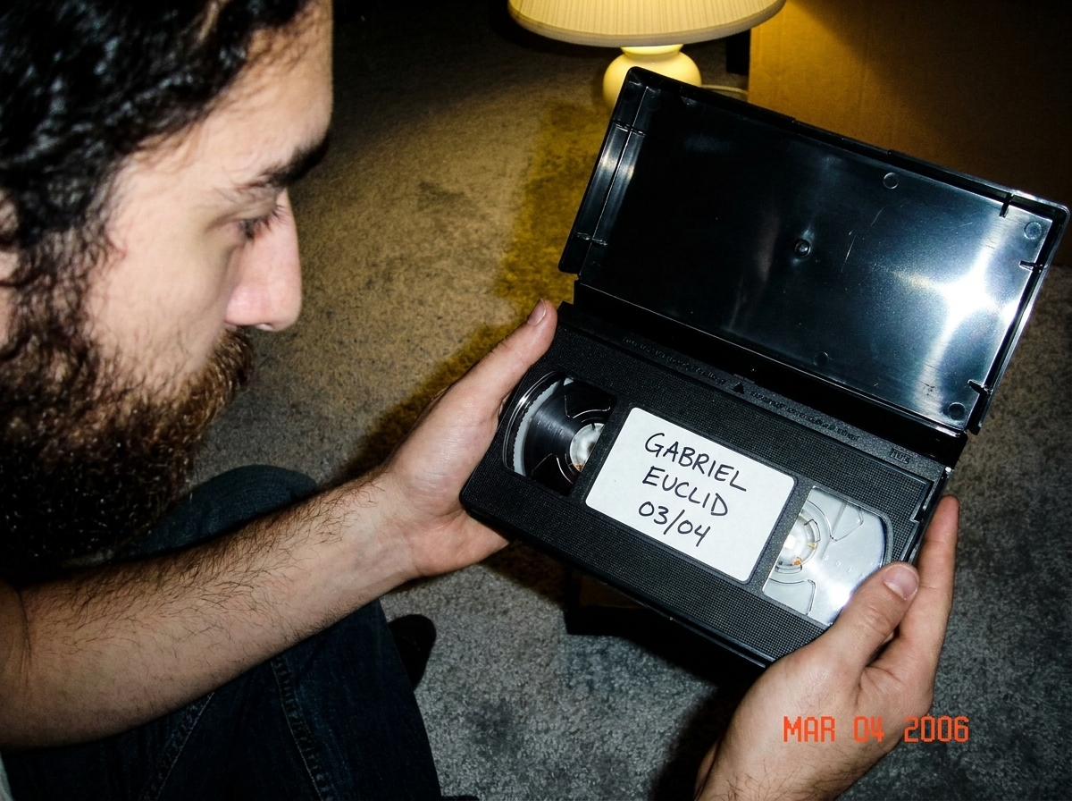 A close-up photo of a VHS tape's paper label, handwritten in black ballpoint pen. The label says 'GABRIEL EUCLID 03/04' in slightly messy handwriting. The tape itself is a generic black clamshell VHS case with no branding visible, held open in someone's hands against a grey carpet background. Late e