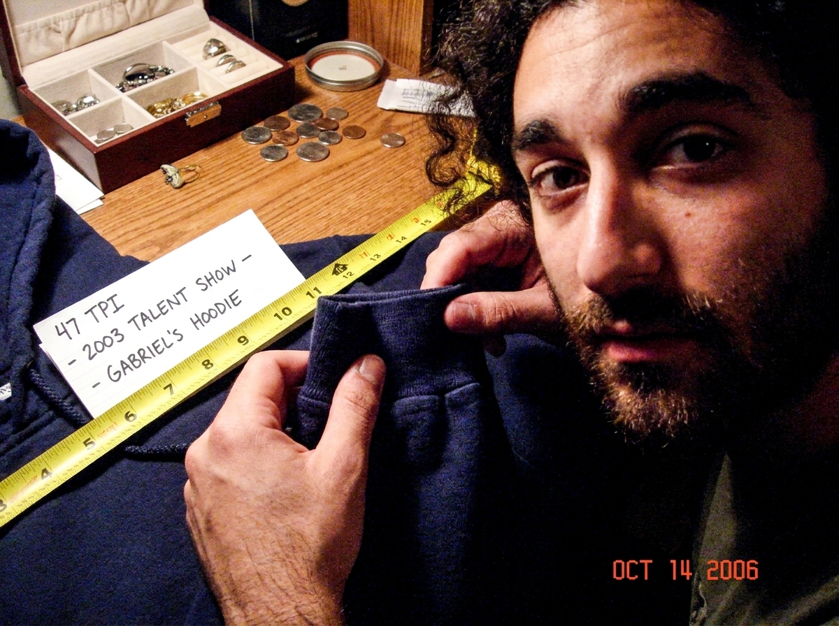 A close-up photo of a dark blue hoodie cuff held in a trembling hand, showing the thread weave. A yellow measuring tape is laid across it, and a piece of white paper with '47 TPI - 2003 TALENT SHOW - GABRIEL'S HOODIE' written in pen is propped next to it. The background is a cluttered wooden desk wi