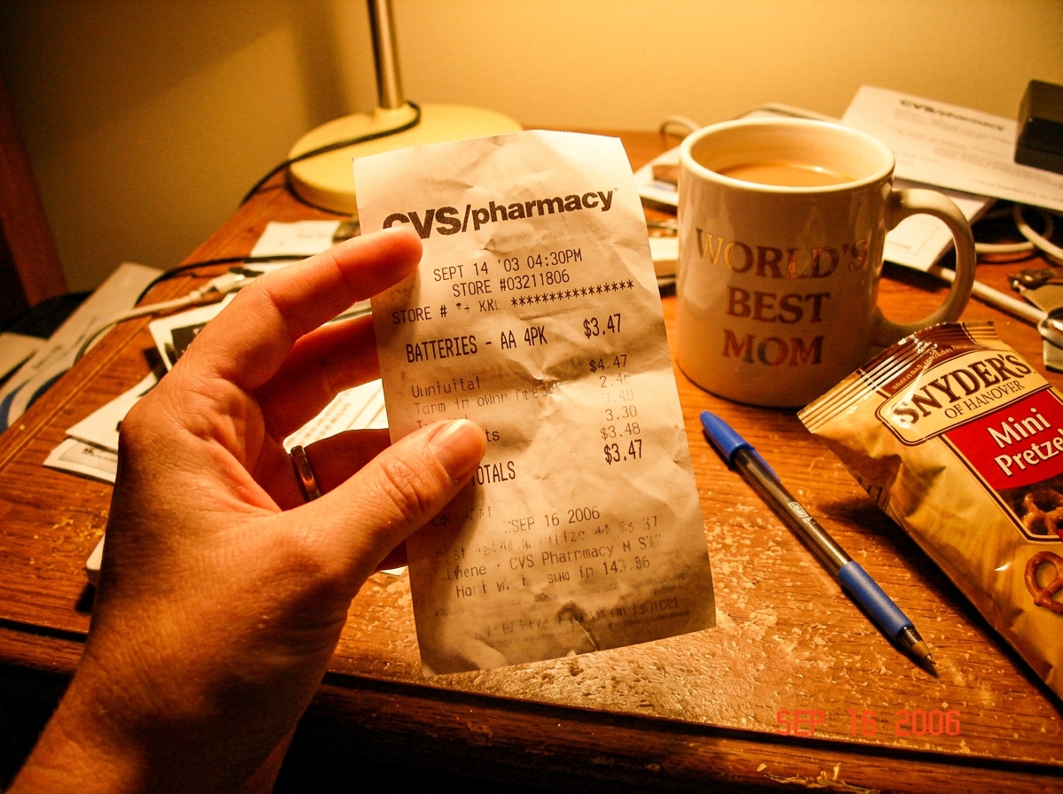 A grainy photo taken on a cheap digital camera in 2006, showing a woman's hand holding a crumpled CVS receipt from September 2003. The receipt is angled toward the camera, with 'BATTERIES - AA 4PK $3.47' visible at the top. The background is a cluttered wooden desk with a ceramic coffee mug that say