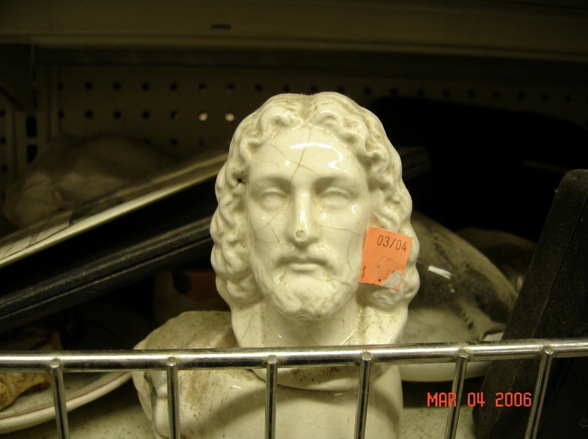 A close-up of a small, cracked white ceramic figurine shaped like a human head sitting on a cluttered thrift store shelf. A faded orange price sticker is visible, marked with handwritten marker: 03/04.