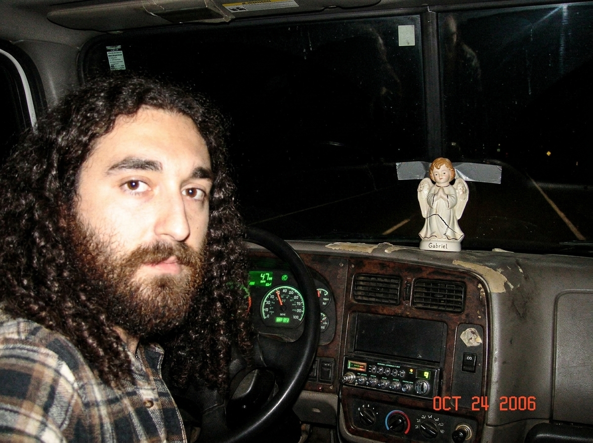 A worn dashboard of a semi truck at night. The speedometer glows faintly green, reading 47 mph. A CB radio sits mounted below, its display lit up. A small ceramic Gabriel figurine is taped to the dash above the radio, a tiny crack running down its face. Beyond the windshield, darkness.