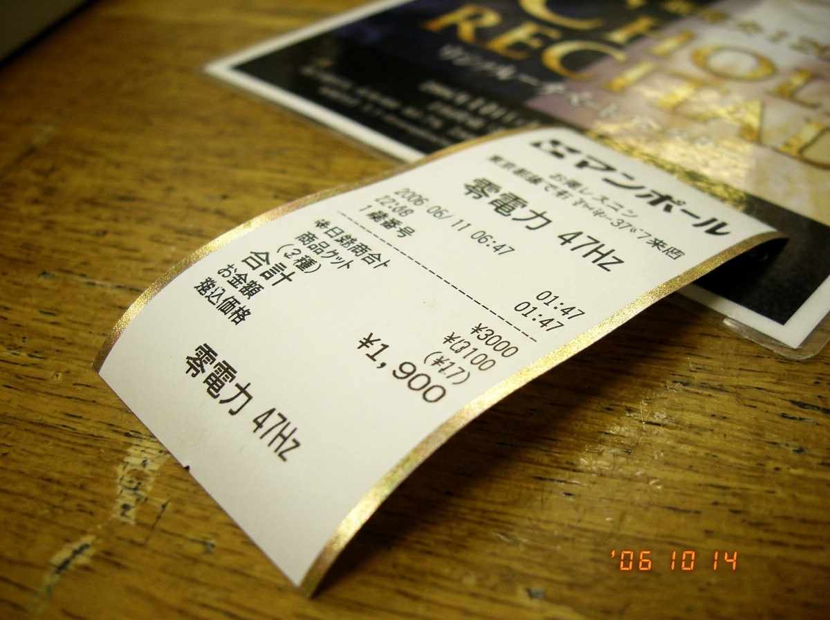 A thermal paper receipt from a Japanese kiosk showing Japanese characters '零電力 47Hz' and a timestamp of 01:47. The paper is slightly curled, with a faint golden shimmer along the edges matching the hue of a nearby recital poster's gold foil lettering. The receipt is lit by warm fluorescent lighting,