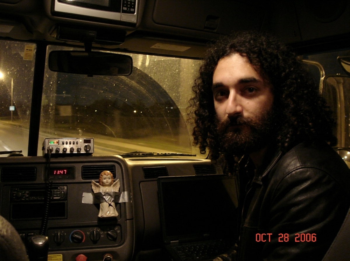 A dark truck cabin at night. The dashboard is dimly lit showing a clock reading 11:47. The CB radio is dark, unlit. A small ceramic Gabriel figurine sits taped to the dash, a crack running down its face. The clock and microwave in the background are both showing blank displays. A laptop screen is da