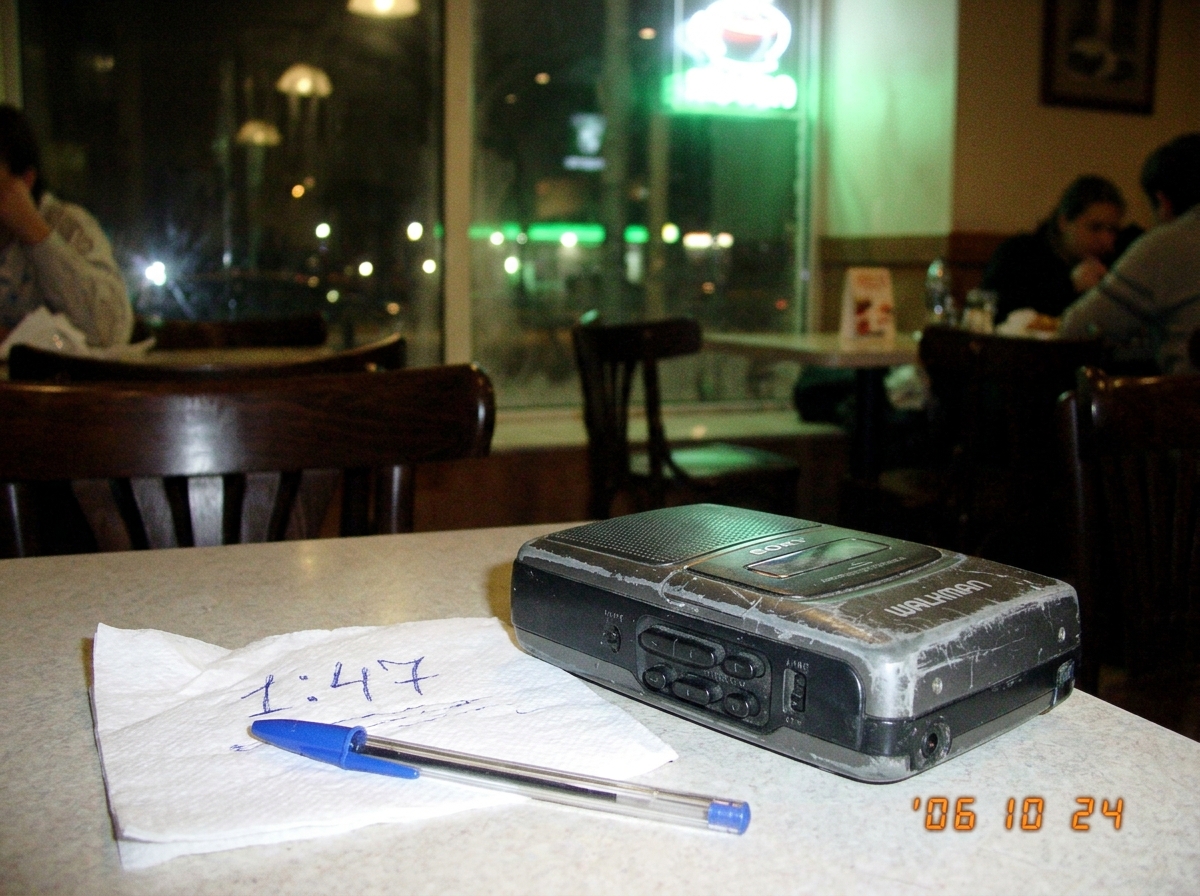 Battered tape recorder on cafe table, greenish glow visible through window, napkin with Cyrillic handwriting '1:47' in foreground