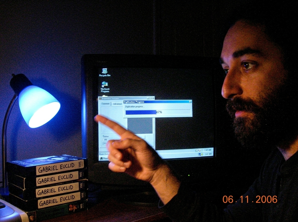 A dark computer desk with a CRT monitor displaying a frozen digitization progress bar at 47% complete. The timestamp on the bottom-right of the Windows 98 taskbar reads 1:47 AM. A desk lamp in the corner casts a blue glow on a stack of VHS tapes labeled 'GABRIEL EUCLID'. A hand is pointing at the mo