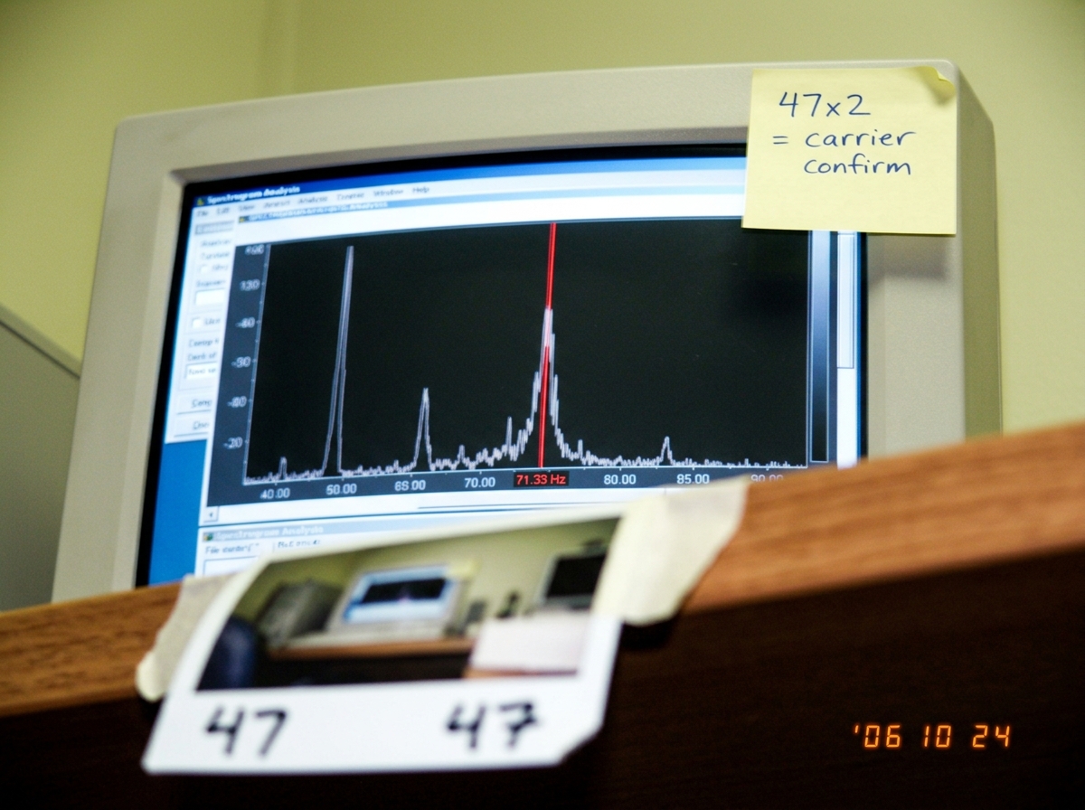 A blurry photo of a computer monitor showing a spectrogram analysis window, a red vertical cursor line highlighting a frequency spike at 71.33 Hz, with a handwritten sticky note on the monitor bezel reading '47x2 = carrier confirm' in blue ballpoint pen. A Polaroid photo is taped to the desk edge, i