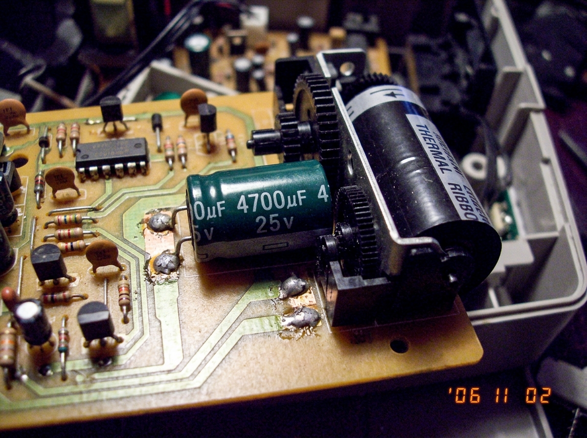 Close-up of a circuit board showing a large cylindrical capacitor labeled '4700uF 25V' next to a thermal printer ribbon mechanism. The board has visible solder joints and traces.