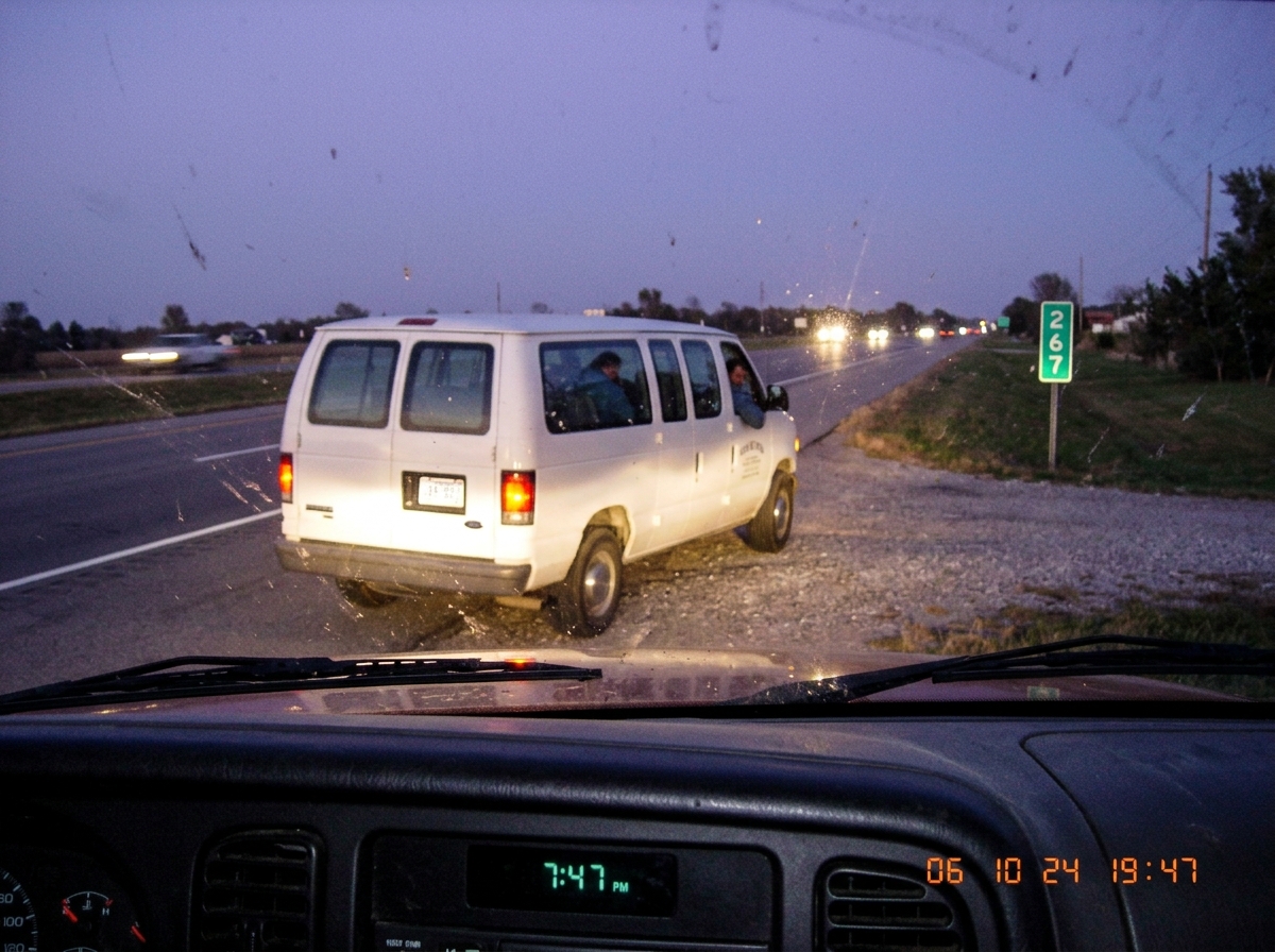 Blurry photo taken from a truck cab through a dusty windshield at dusk. A white church van is pulling out of a gravel lot near a highway mile marker 267 sign. The van's license plate is visible but grain makes it hard to read. Through the van's open driver window, the silhouette of a man in a denim 