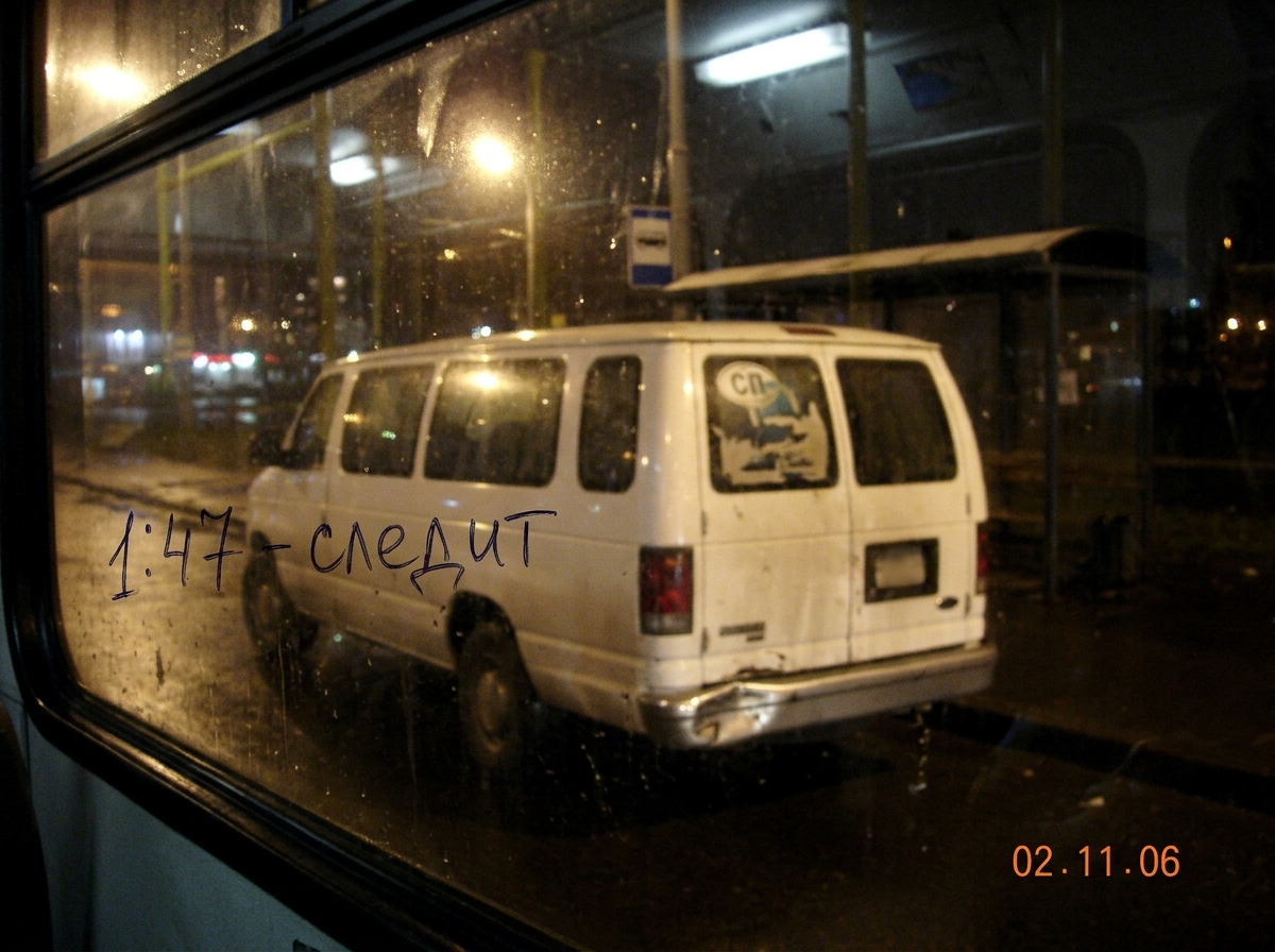 A blurry photo taken from inside a bus window at night, showing a white passenger van parked at a bus stop. The van has a dented rear bumper and a faded sticker on the back window. A handwritten note on the photo in Cyrillic says '1:47 - slezhit' meaning 'watches/wait'. The photo quality is poor, ta