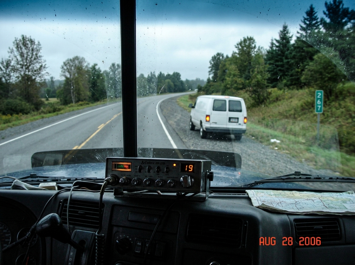 Blurry photo taken through a semi truck windshield. A white cargo van is parked on the shoulder of a two-lane highway. A mile marker sign reading '267' is partly visible in the background. The dashboard of the truck shows a CB radio unit mounted below the windshield, display showing channel 19. Date