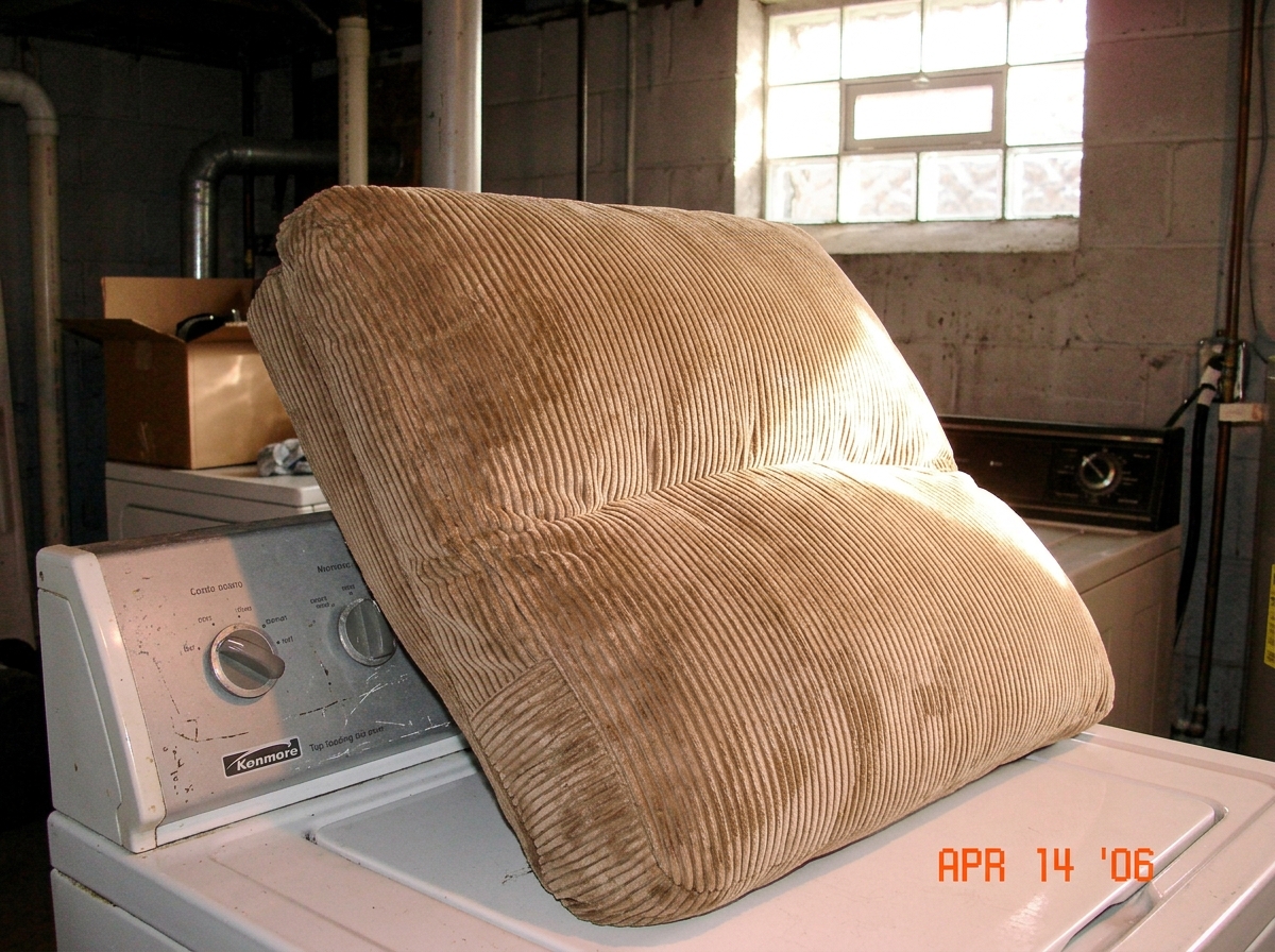 A tan corduroy recliner cushion leaning against a dryer, freshly washed and damp, with sunlight hitting the fabric through a basement window.