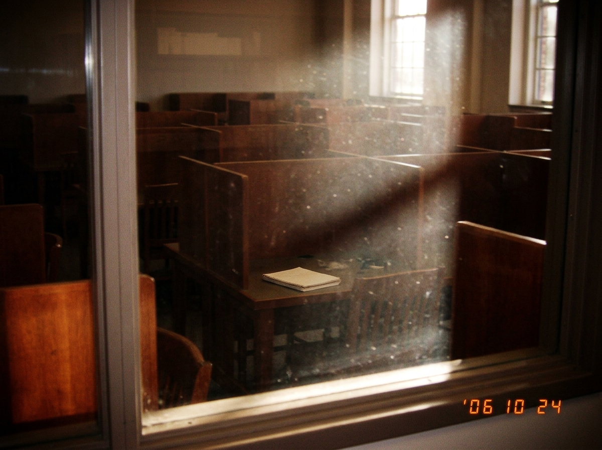 A hazy photo through a library window: rows of empty study carrels in late afternoon light, dust motes suspended in a sunbeam, one open notebook left on a table with nothing written on the visible page