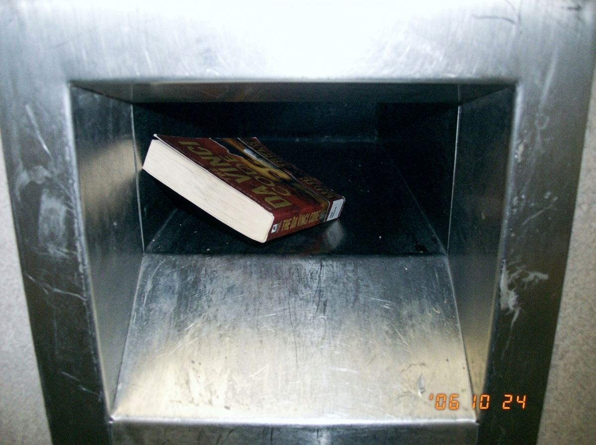 a blurry photo of a library book return slot taken from above, looking down into the dark slot, a book halfway in, the metal is worn silver