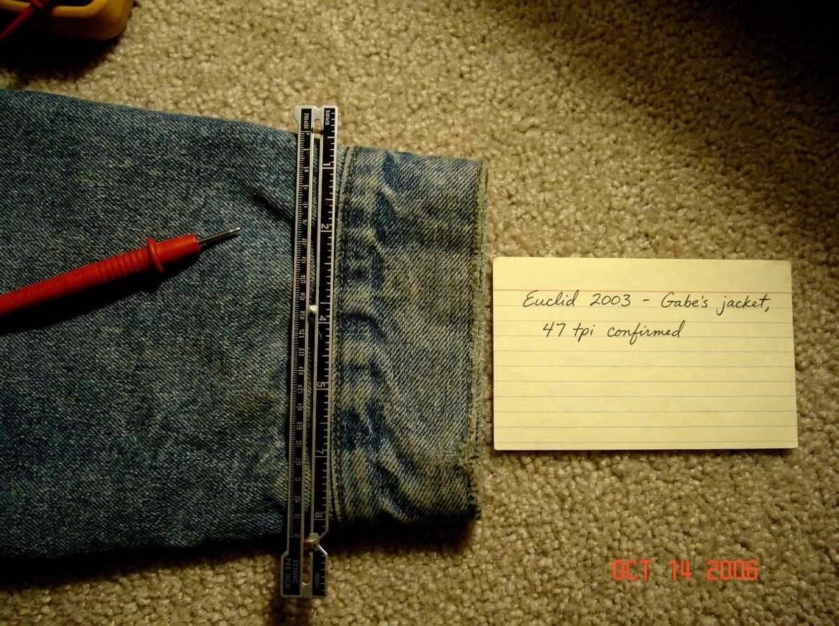 A close-up photo of a faded denim jacket cuff laid flat on a beige carpet next to a sewing gauge. The cuff shows visible weave grain at roughly 45 threads per inch counted against the gauge. A small yellowed index card in the corner reads 'Euclid 2003 - Gabe's jacket, 47 tpi confirmed' in cursive ha