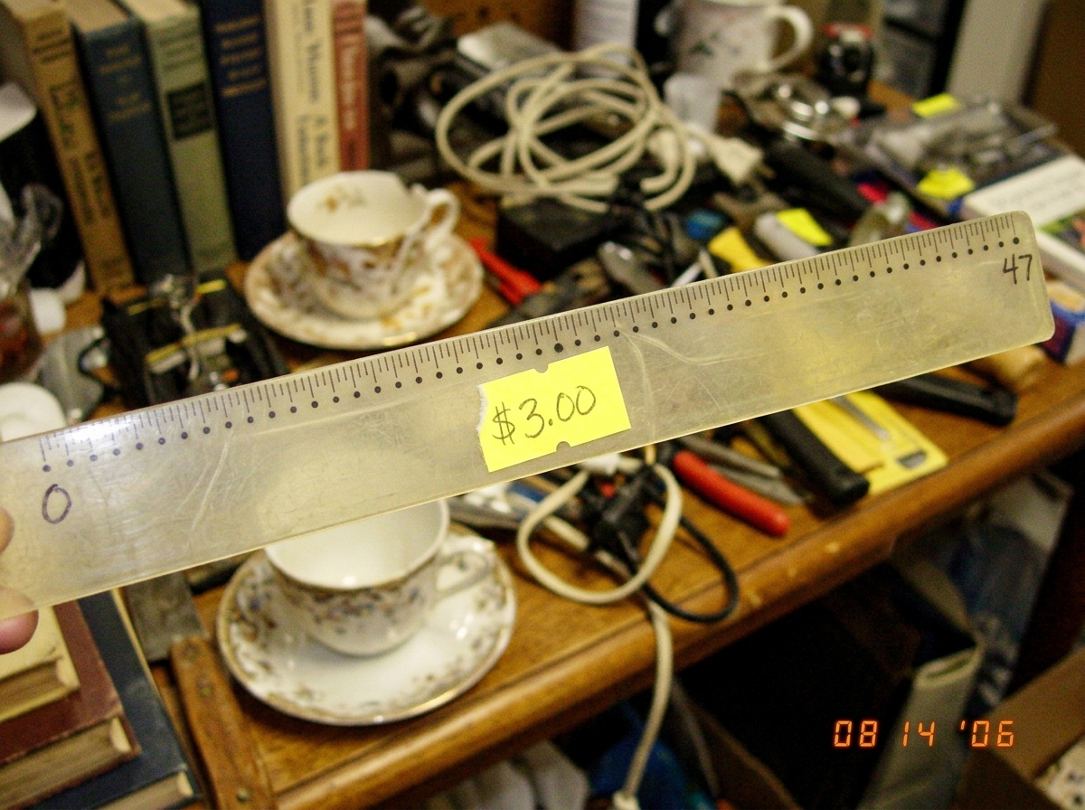 A blurry photo of a cheap plastic 12-inch ruler with tiny dots marking 47 equal divisions instead of the usual 16ths or 8ths. The ruler is slightly warped and has a yellow price sticker reading $3.00. In the background, a cluttered yard sale table with other junk like old books and a chipped teacup.