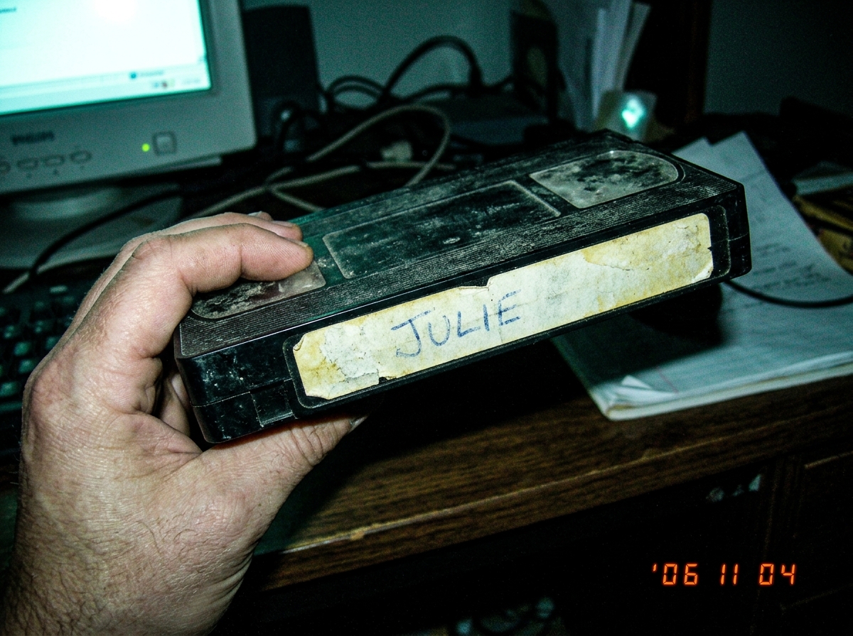 A close up photo of a dusty black VHS tape held in a hand, the spine label is white and yellowed with age, the word JULIE is handwritten in faded blue ink, the lighting is dim and green tinted from a computer monitor