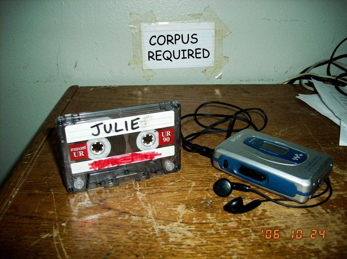 A blank cassette tape labeled 'JULIE' with a red marker smudge where the tracklist should be, sitting on a wooden desk next to a disconnected Walkman. The desk has a single index card taped to the wall reading 'CORPUS REQUIRED' in Comic Sans MS.