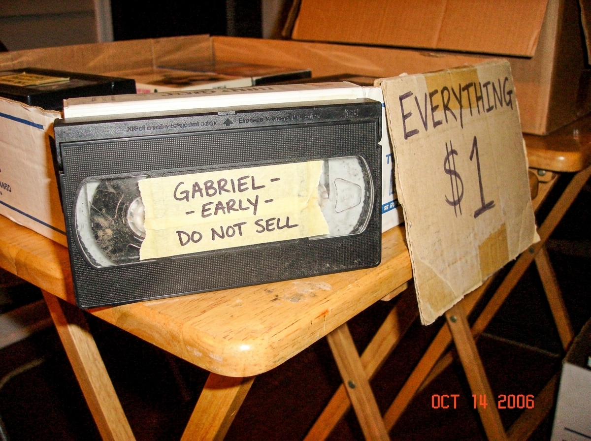 A blurry camera phone photo of a dusty VHS tape sitting on a folding table at a yard sale. The tape has a handwritten 'GABRIEL - EARLY - DO NOT SELL' note stuck to it with yellowed tape. No other markings visible. The table has a faded 'EVERYTHING $1' sign.