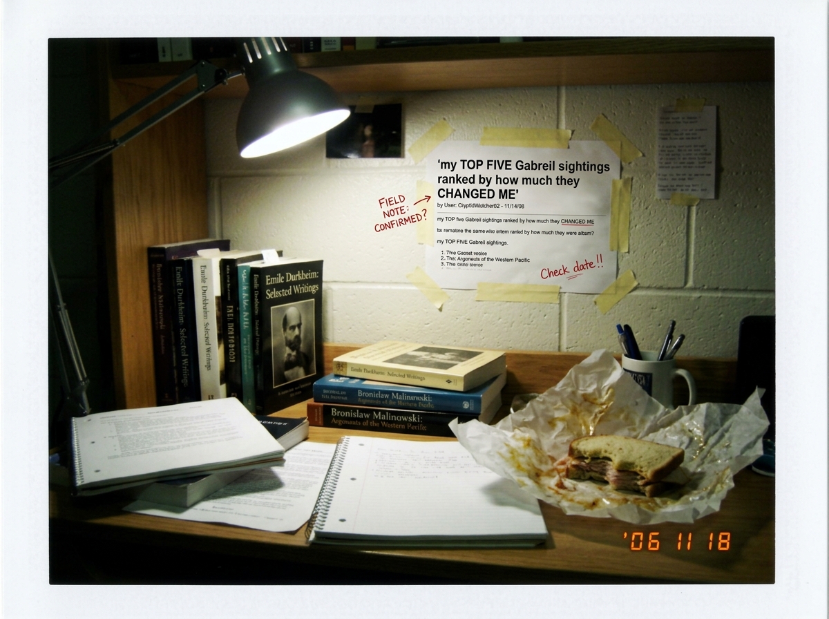 A Polaroid photo of a college dorm room desk cluttered with anthropology textbooks (Durkheim, Malinowski), a half-eaten turkey sandwich on wax paper, and a printed screenshot of the forum thread title 'my TOP FIVE Gabreil sightings ranked by how much they CHANGED ME' taped to the wall like a field n