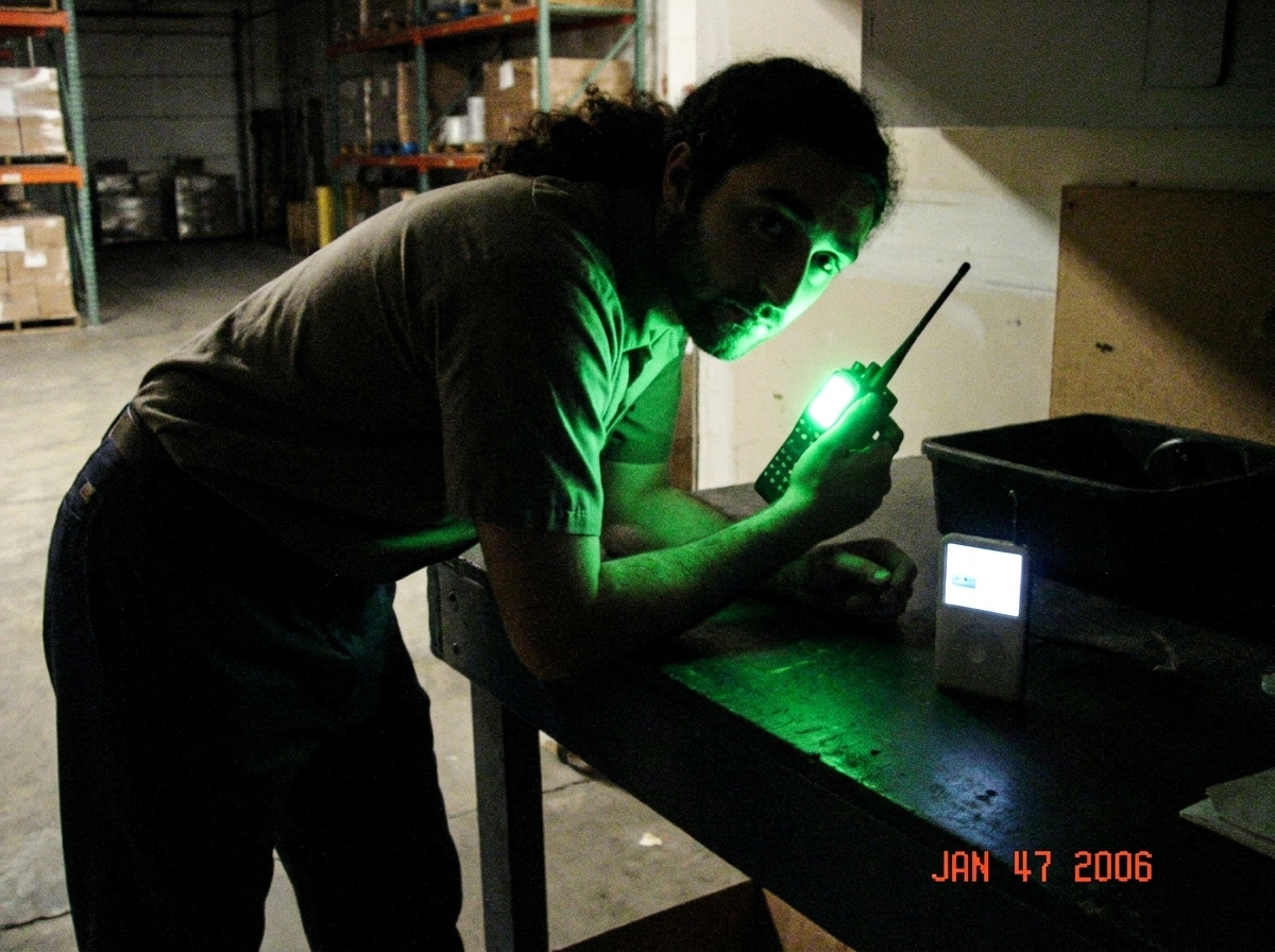Photo of my walkie talkie green glow on warehouse table at 1:47, shadows strong like Gabriel blessing, uniform pants beside MP3 player full power
