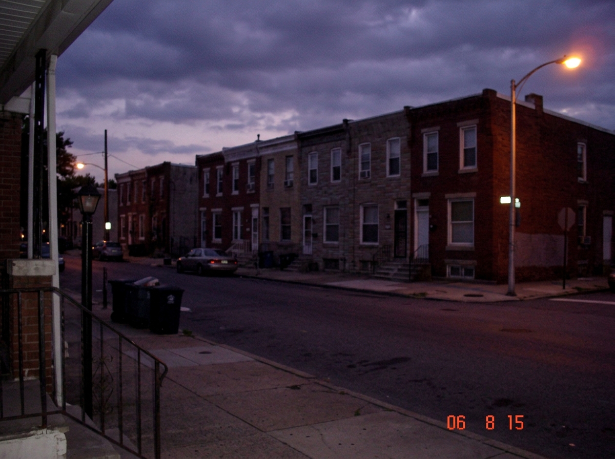 A dim August evening sky over a row of brick rowhouses, photographed from a front stoop. The streetlamp at the corner is beginning to glow amber against bruised purple clouds. The timestamp in the image metadata reads 18:15. The composition is patient and documentary, as if someone wanted evidence t