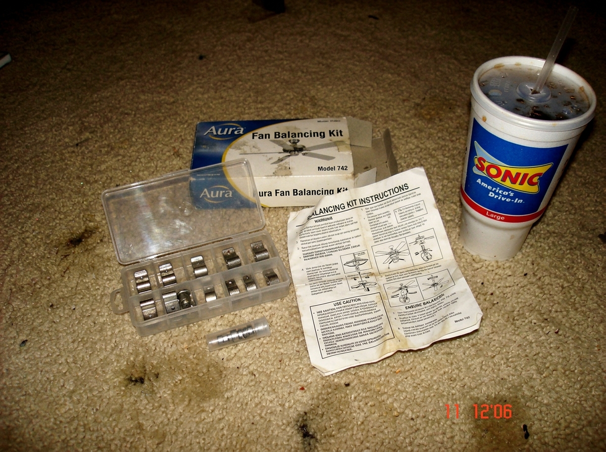 A blurry camera phone photo of a ceiling fan balancing kit spread out on a beige carpet floor next to a Sonic drive-in cup. The kit includes small metal clips, a plastic case, and a folded instruction manual that looks aggressively unhelpful.