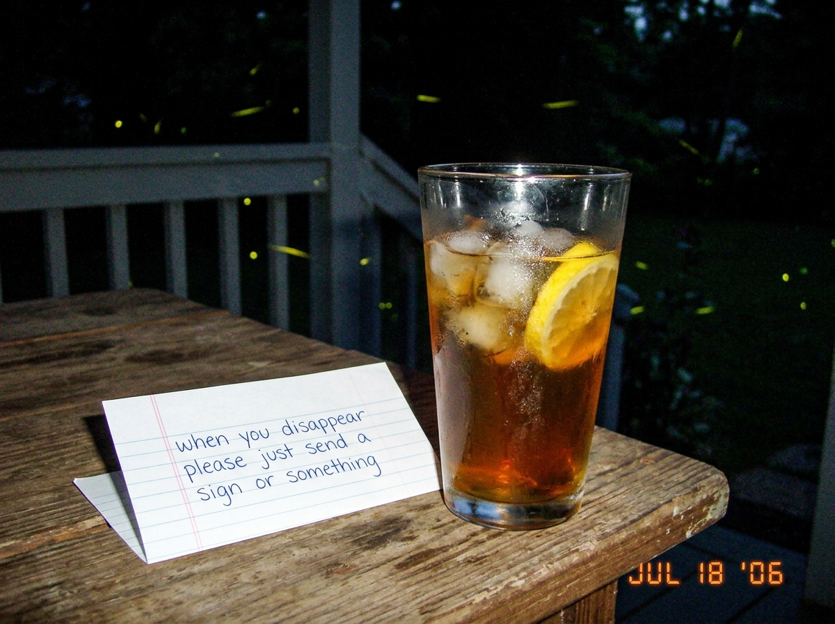 A glass of iced tea on a wooden porch table with fireflies floating in the background and a handwritten note that says 'when you disappear please just send a sign or something'