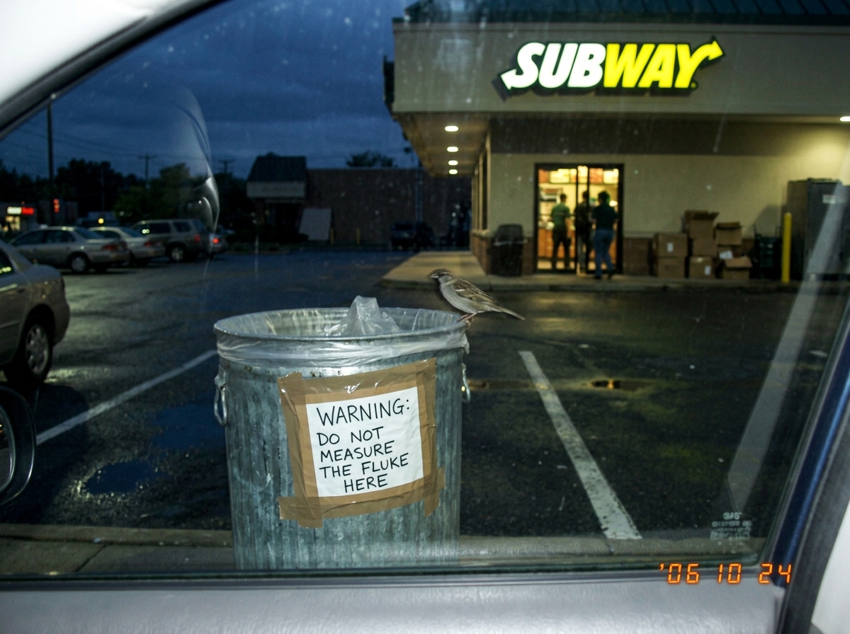 A blurry cellphone photo taken through a car window at dusk, showing a parking lot behind a sandwich shop. A dented trash can with a hand-written sign taped to it reading 'WARNING: DO NOT MEASURE THE FLUKE HERE' in sharpie. A single bird is perched on the rim of the trash can.