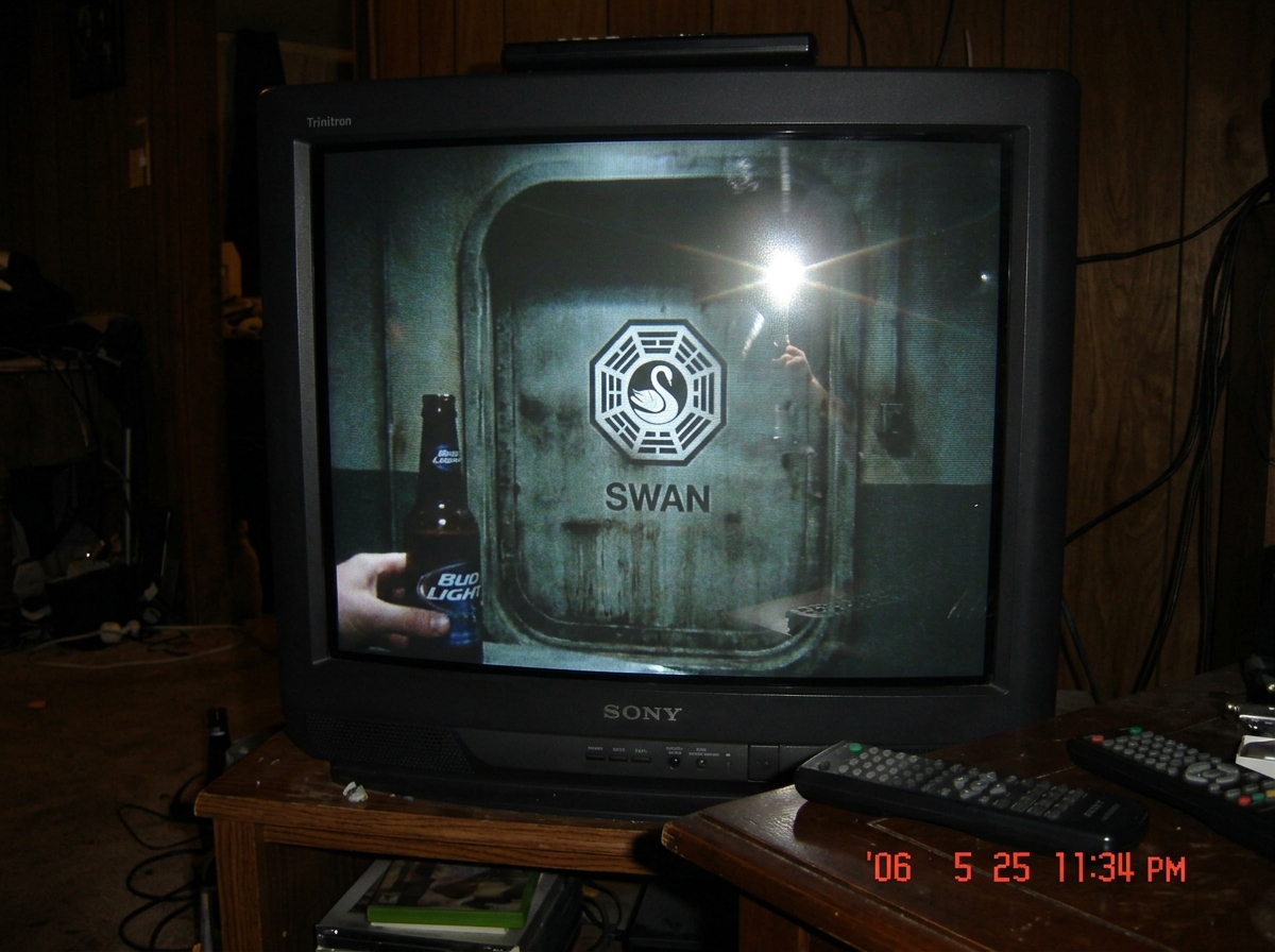 A blurry CRT television screen showing the last scene of the Lost season 2 finale from 2006 - the hatch door closing, grainy freeze-frame, with a beer bottle reflection in the glass and a remote control visible on the table edge