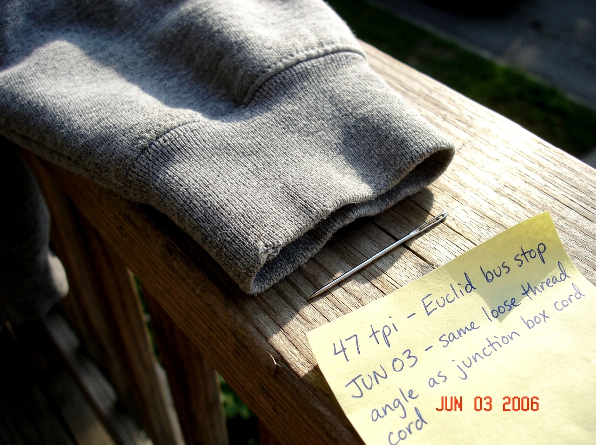 A close-up photograph of a worn gray hoodie cuff lying on a wooden porch railing in late afternoon light. The weave is clearly visible with individual threads countable by eye. A silver sewing needle rests next to the fabric for scale. A yellow sticky note in the corner has handwriting that says '47