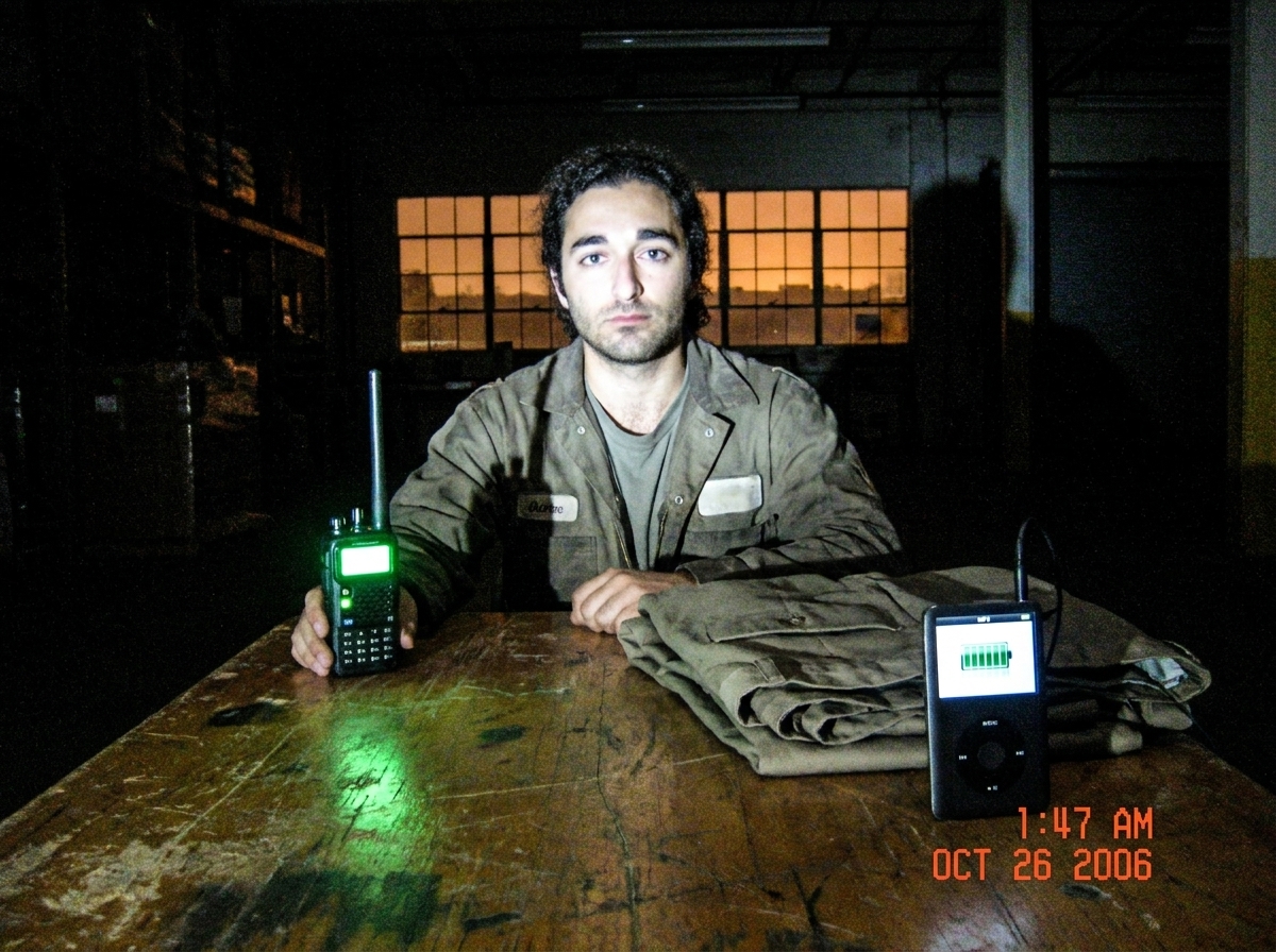 Photo of a green-glow walkie talkie on a warehouse table at 1:47 AM, shadows strong like Gabriel's blessing, my uniform pants laid out beside the MP3 player with full power. The warehouse district window shows the orange sky from earlier tonight.